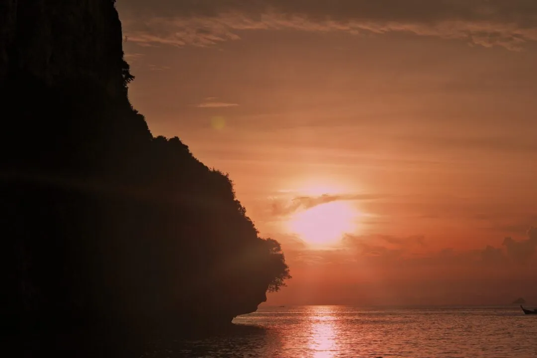 Sunlight breaking around a jungle-topped limestone cliff with a small boat below, Thailand.
