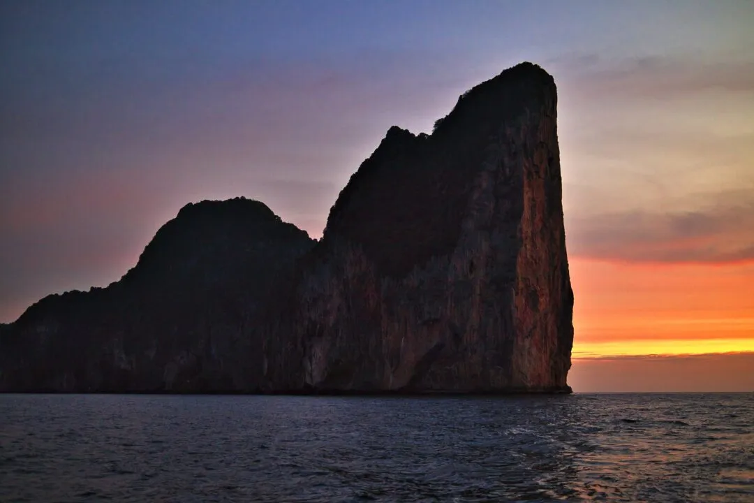 A jagged limestone pinnacle island silhouetted against a warm sunset sky in Thailand.