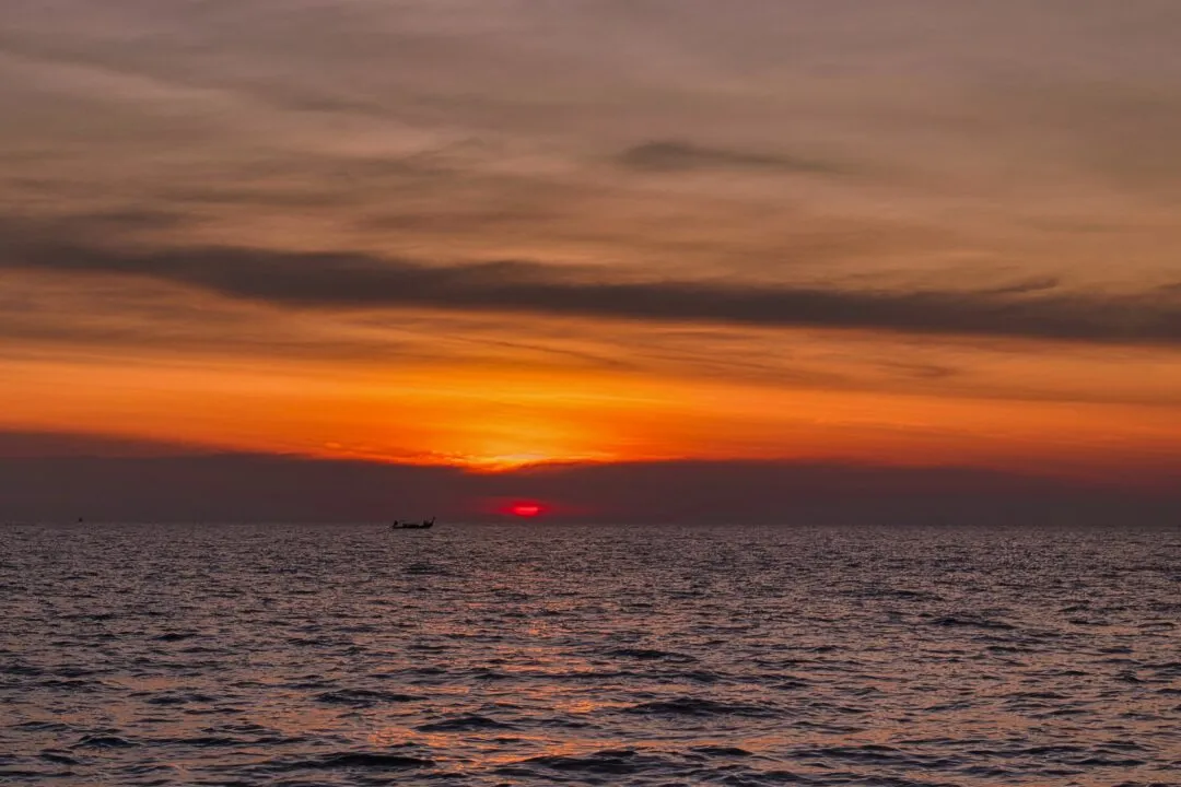 A lone fishing boat silhouetted against a glowing red-orange sunset sky in Thailand.