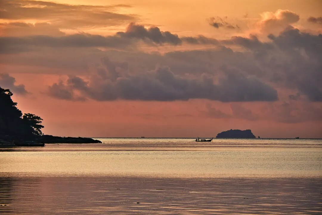 A longtail boat silhouetted against a hazy island at dusk on the Thai coast.