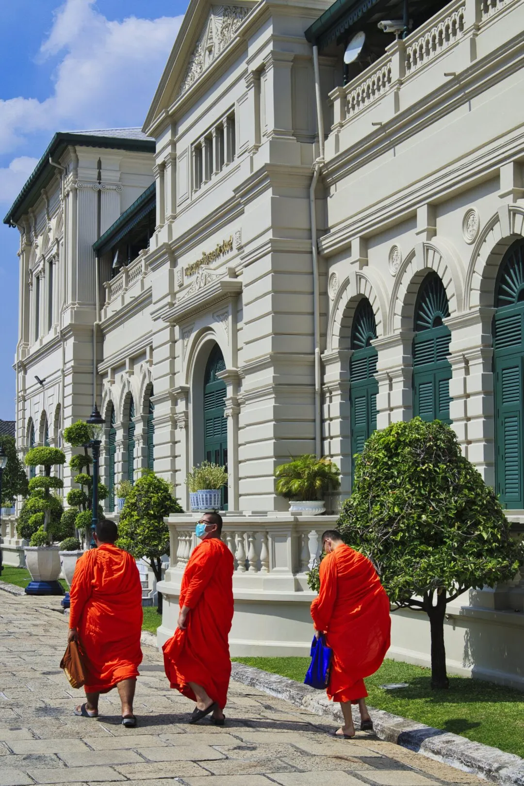 Three monks in vivid orange robes walking past a colonial-style facade in Bangkok.