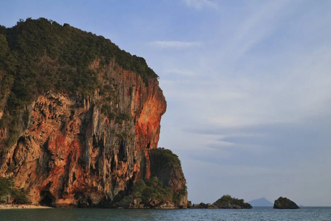 A large limestone cliff face streaked with rust-orange mineral bands above a beach, Thailand.
