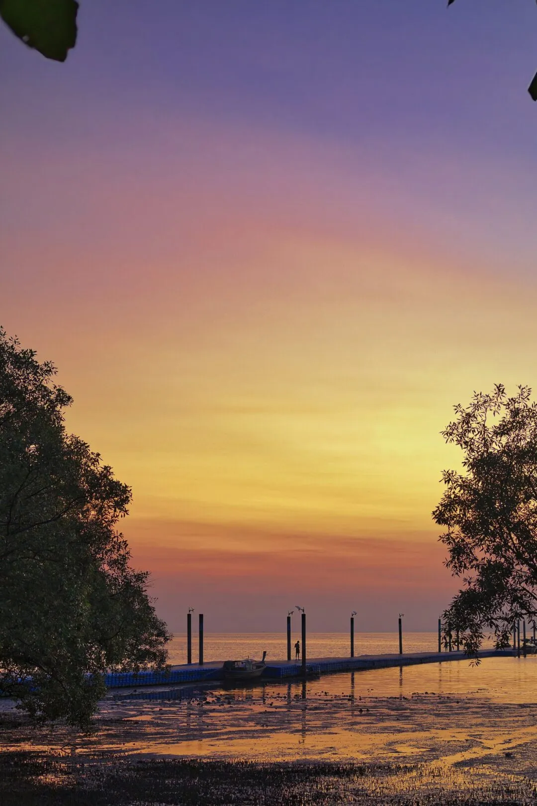 A wooden pier over calm tidal water at dusk, with a figure and a moored boat, Thailand.
