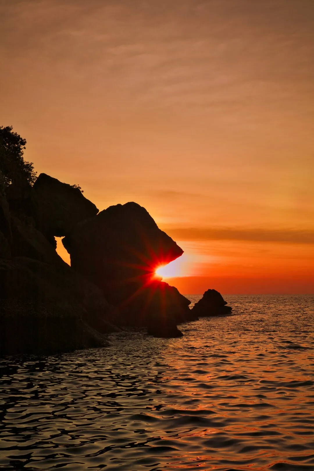A sea-arch rock formation silhouetted against a sunburst on the Thai coast.