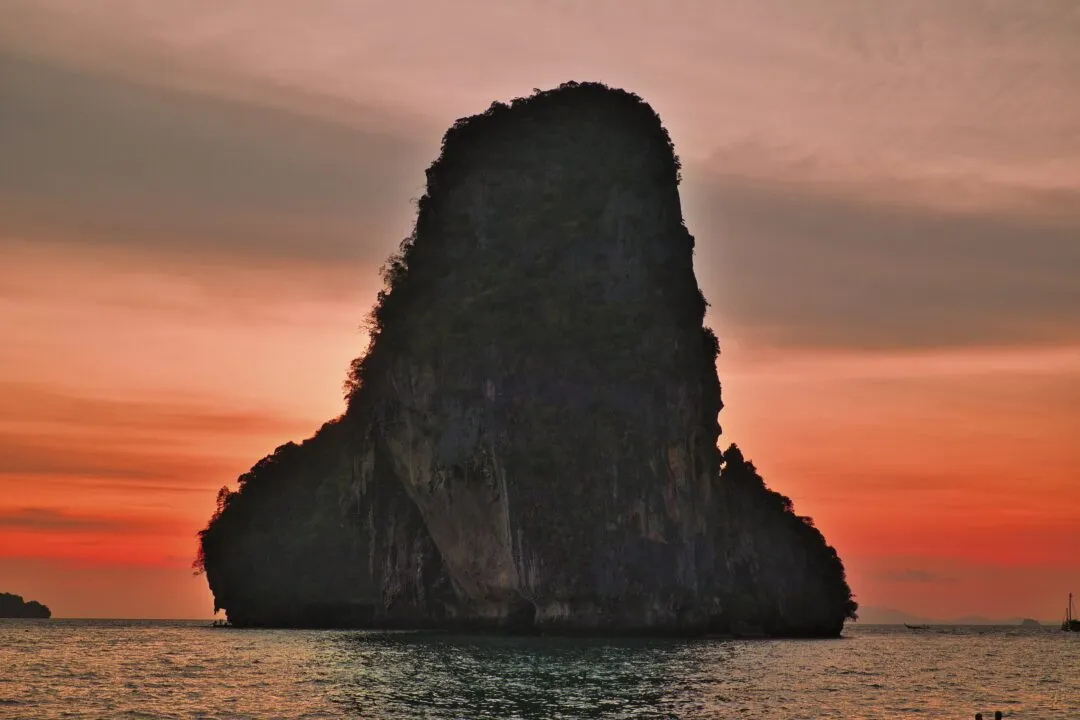 A large sea-stack island silhouetted at sunset with calm reflective water, Thailand.