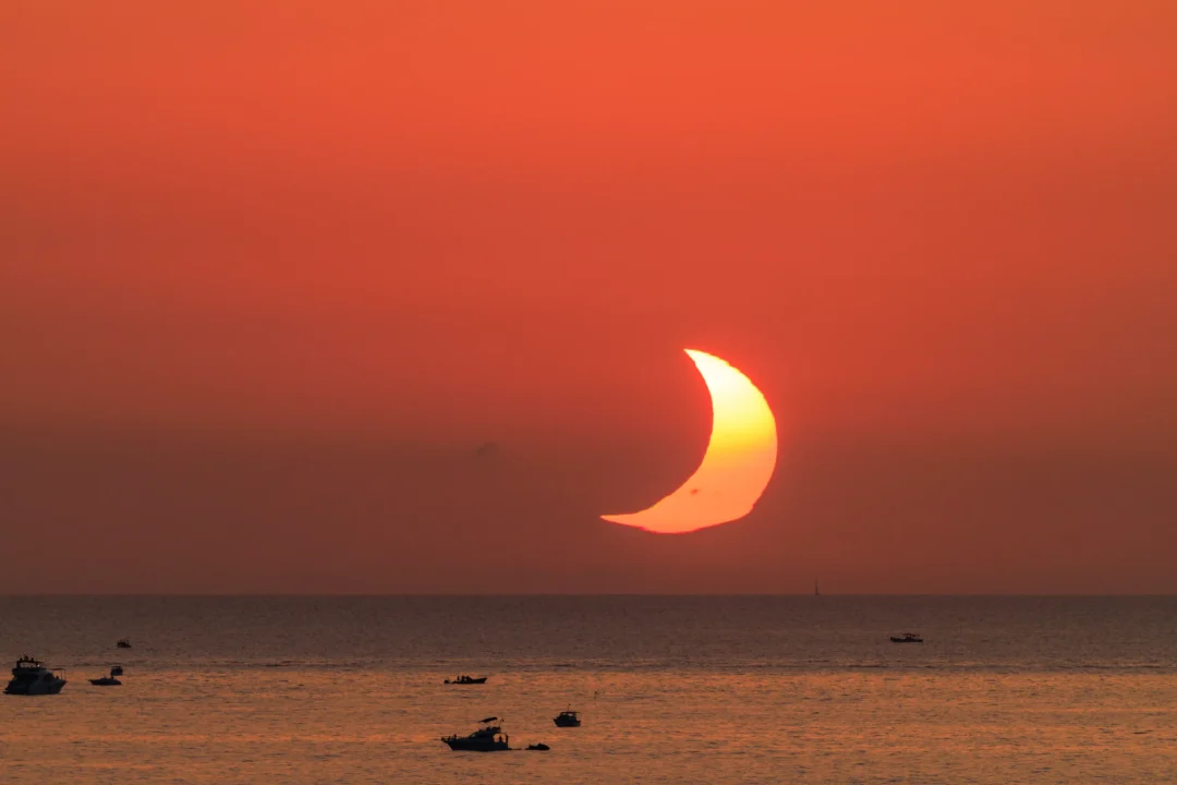 The crescent shaped eclipsed sun touching the sea horizon off Ibiza, with small boats silhouetted on the water