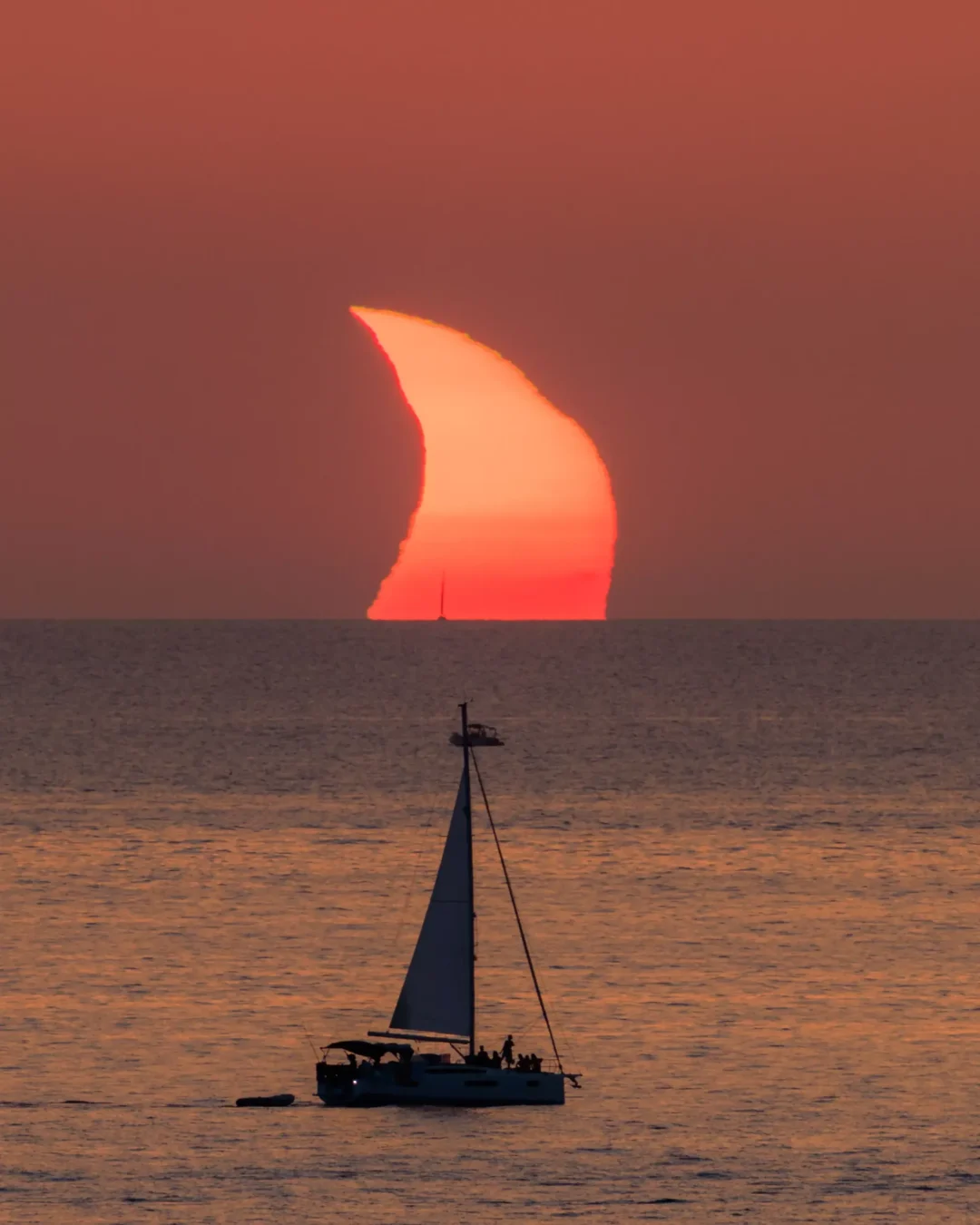 Portrait view of a crescent eclipsed sun setting into the sea above a silhouetted sailboat off the coast of Ibiza