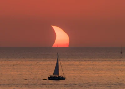A thin crescent of the eclipsed sun setting into the sea behind the silhouette of a sailboat off Ibiza