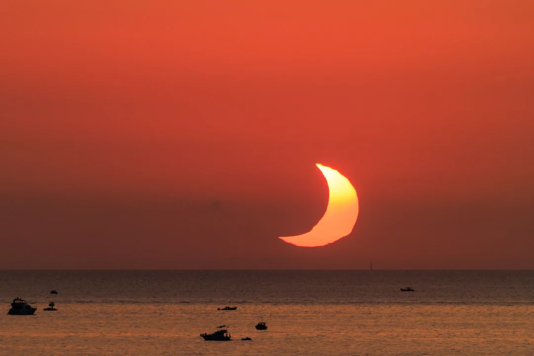 The eclipsed crescent sun partially submerged below the sea horizon off the coast of Ibiza, boats silhouetted on the water