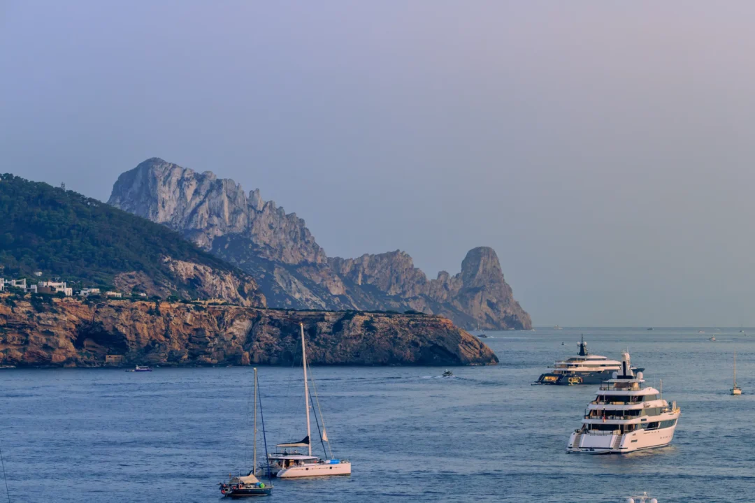 The rocky silhouette of Es Vedrà island near Ibiza during blue hour, with anchored yachts and sailboats in the foreground