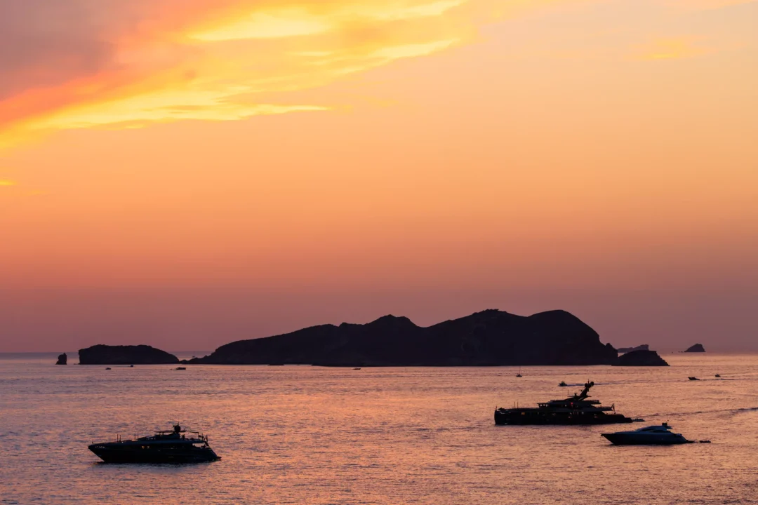 Twilight sky over the island of Es Vedrà near Ibiza after sunset, with yachts anchored in the calm water below