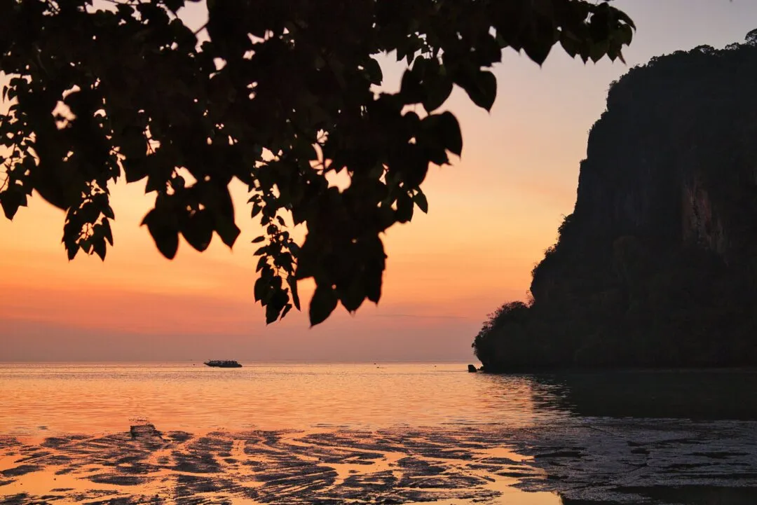 A sunset over calm sea framed by foliage and a limestone cliff, with colour-streaked wet sand, Thailand.