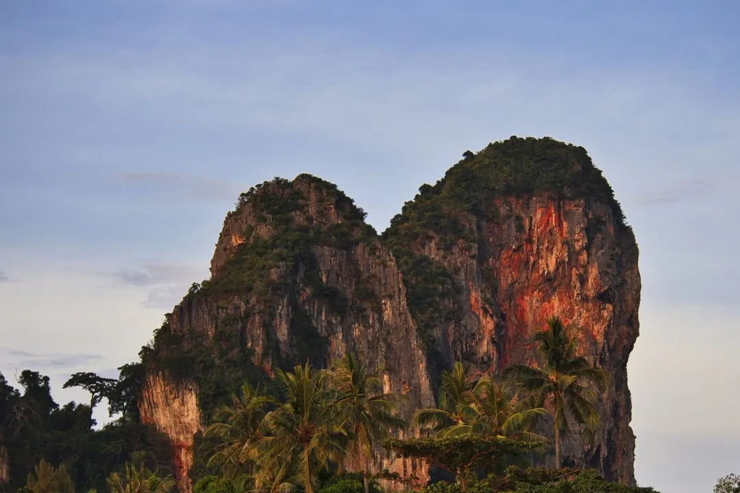 A twin-peaked limestone karst tower rising above coconut palms in Thailand.