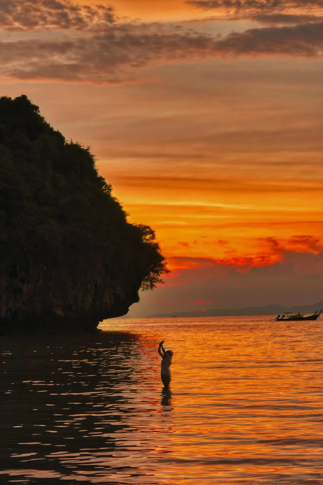 A person silhouetted wading in shallow sea water at sunset beside a limestone cliff in Thailand.