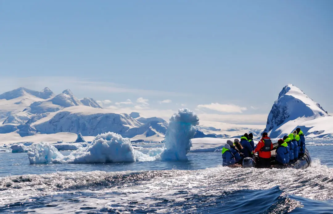 Small zodiac boat dwarfed beneath a colossal sculpted iceberg and mountain wall, Antarctica
